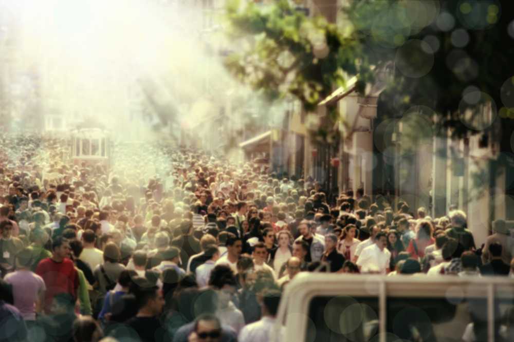 Eine Menschenmenge auf der Straße, die einzelnen Gesichter sind nicht zu erkennen. Im Hintergrund sind Bäume und grelles Sonnenlicht.